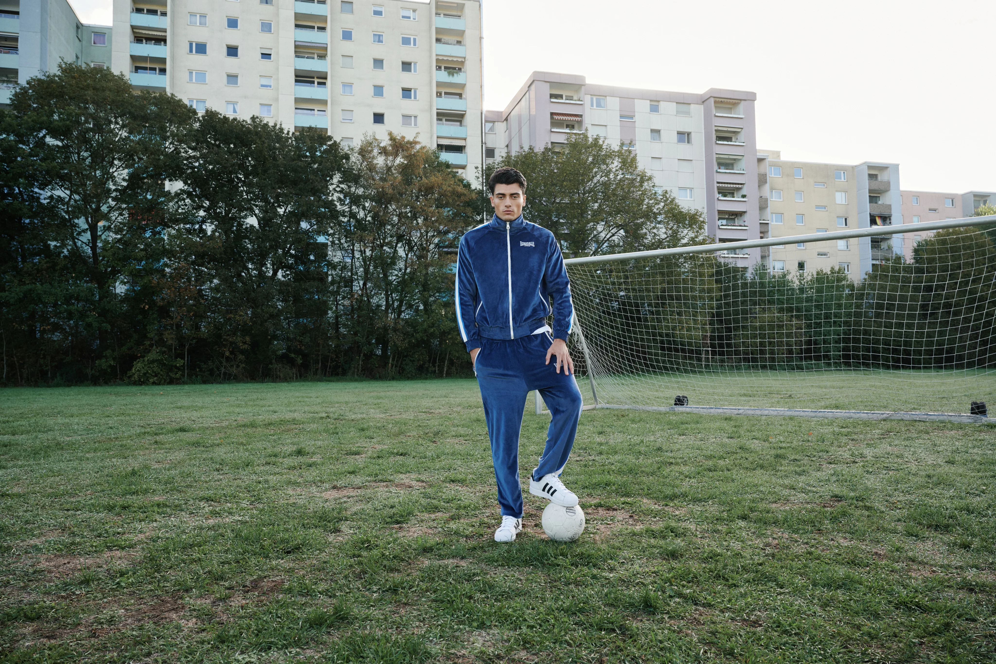 Man in a blue Lonsdale tracksuit with one foot on a football in front of a goal, apartment blocks behind