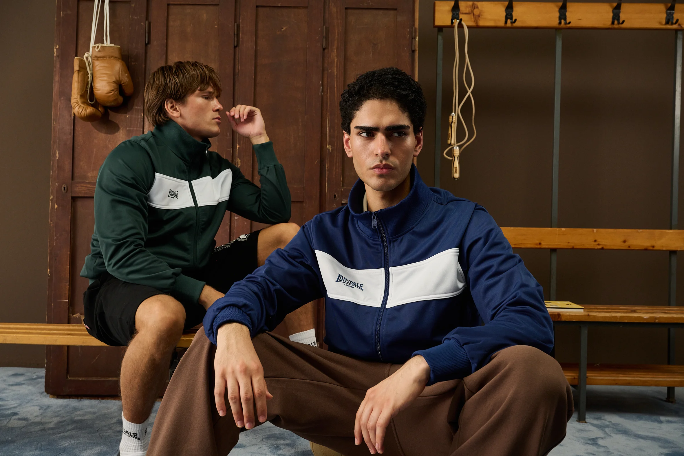 Two men in Lonsdale track jackets on a bench in an old locker room