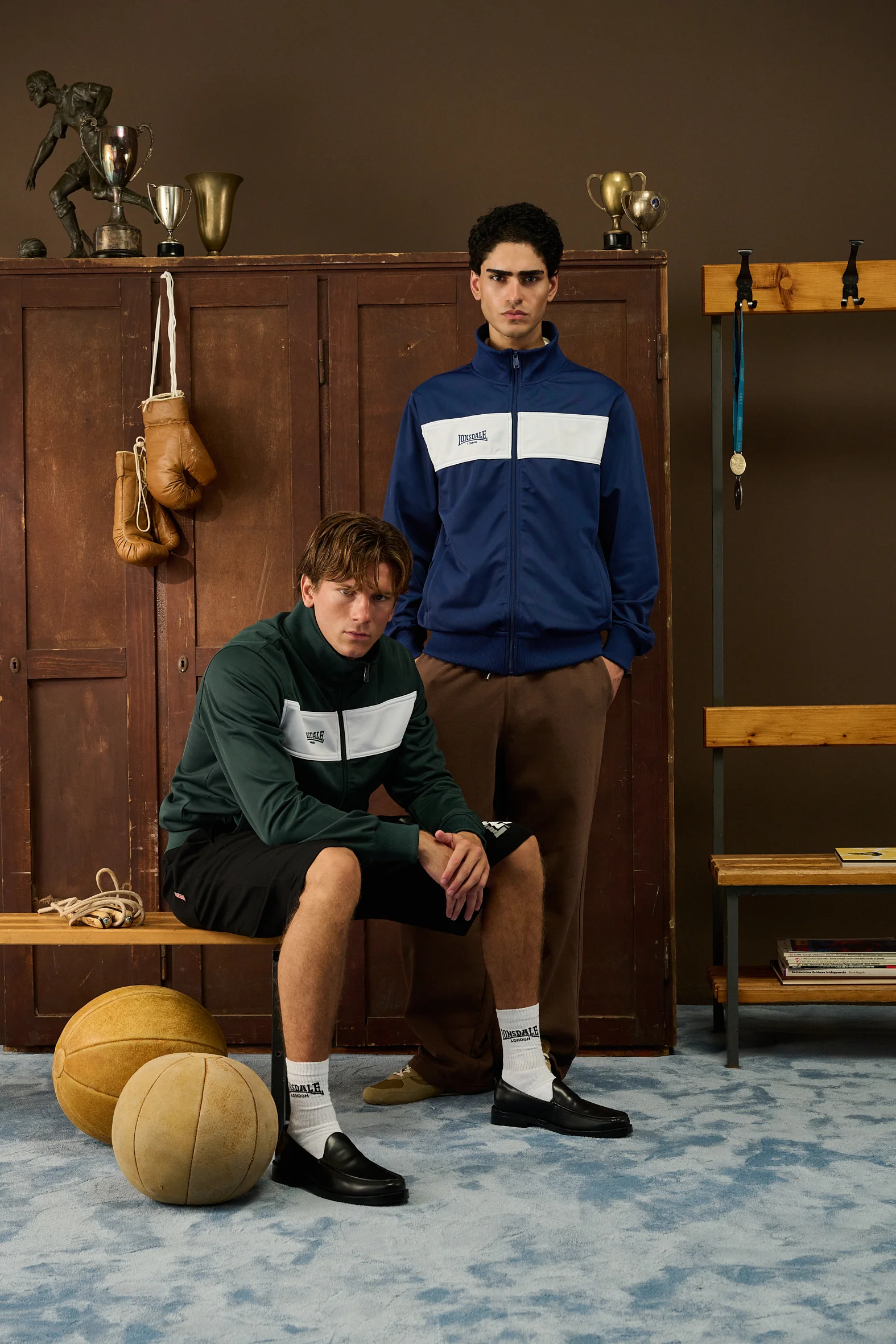 Two men in Lonsdale track jackets in front of wooden lockers, boxing gloves on a hook
