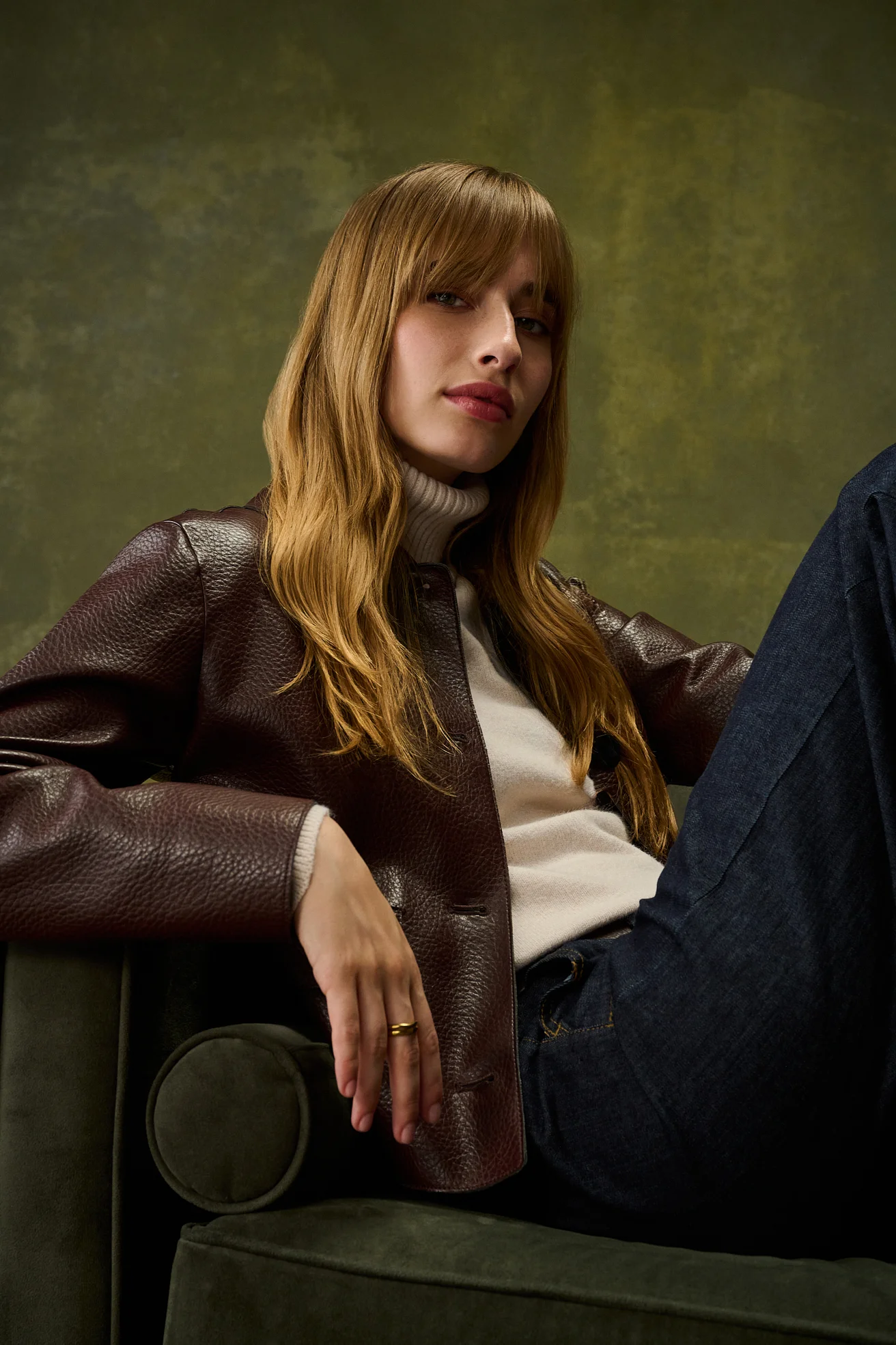 Model in a brown leather jacket and jeans leaning on the arm of a green velvet sofa