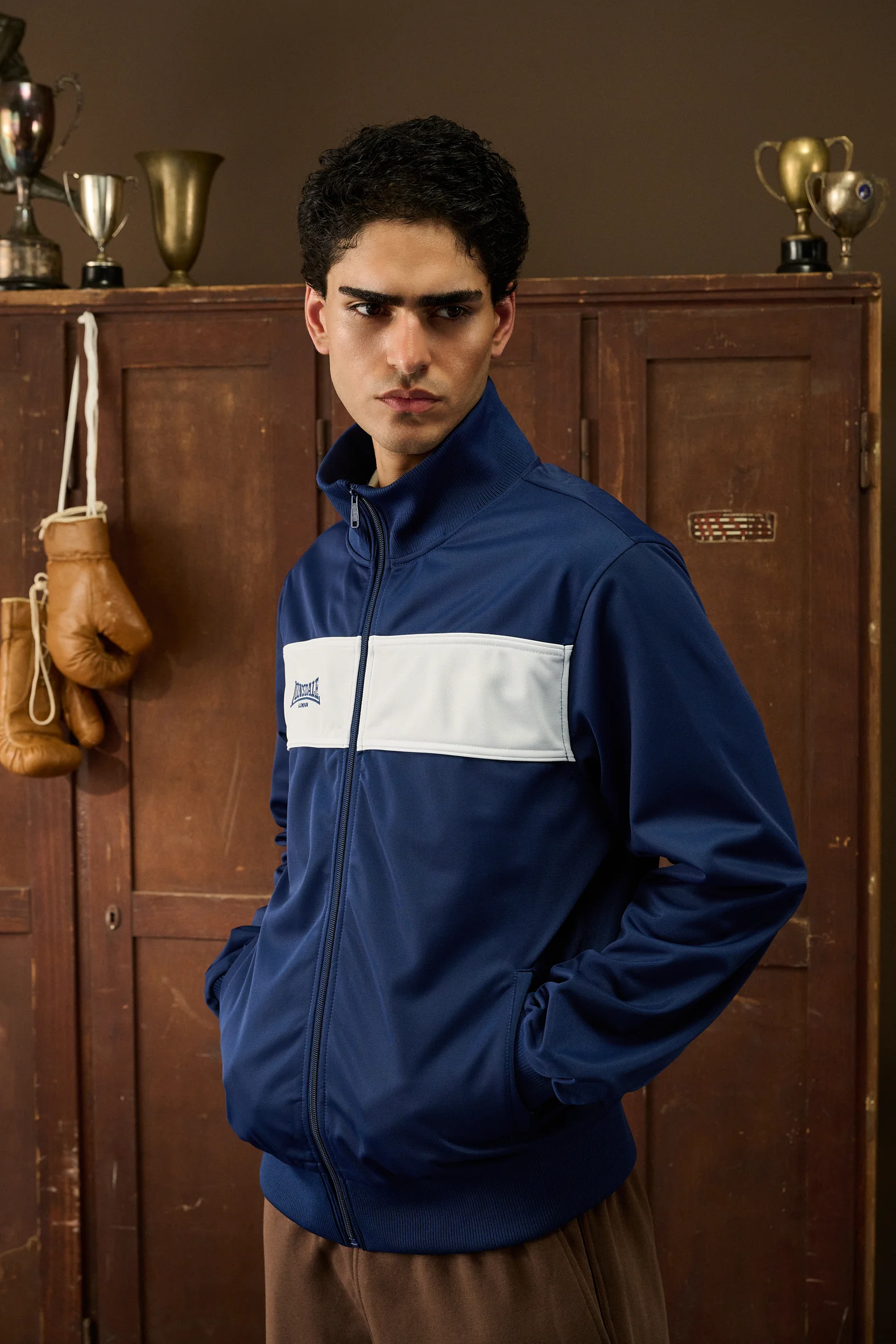 Man in a blue Lonsdale track jacket in front of wooden lockers and trophies