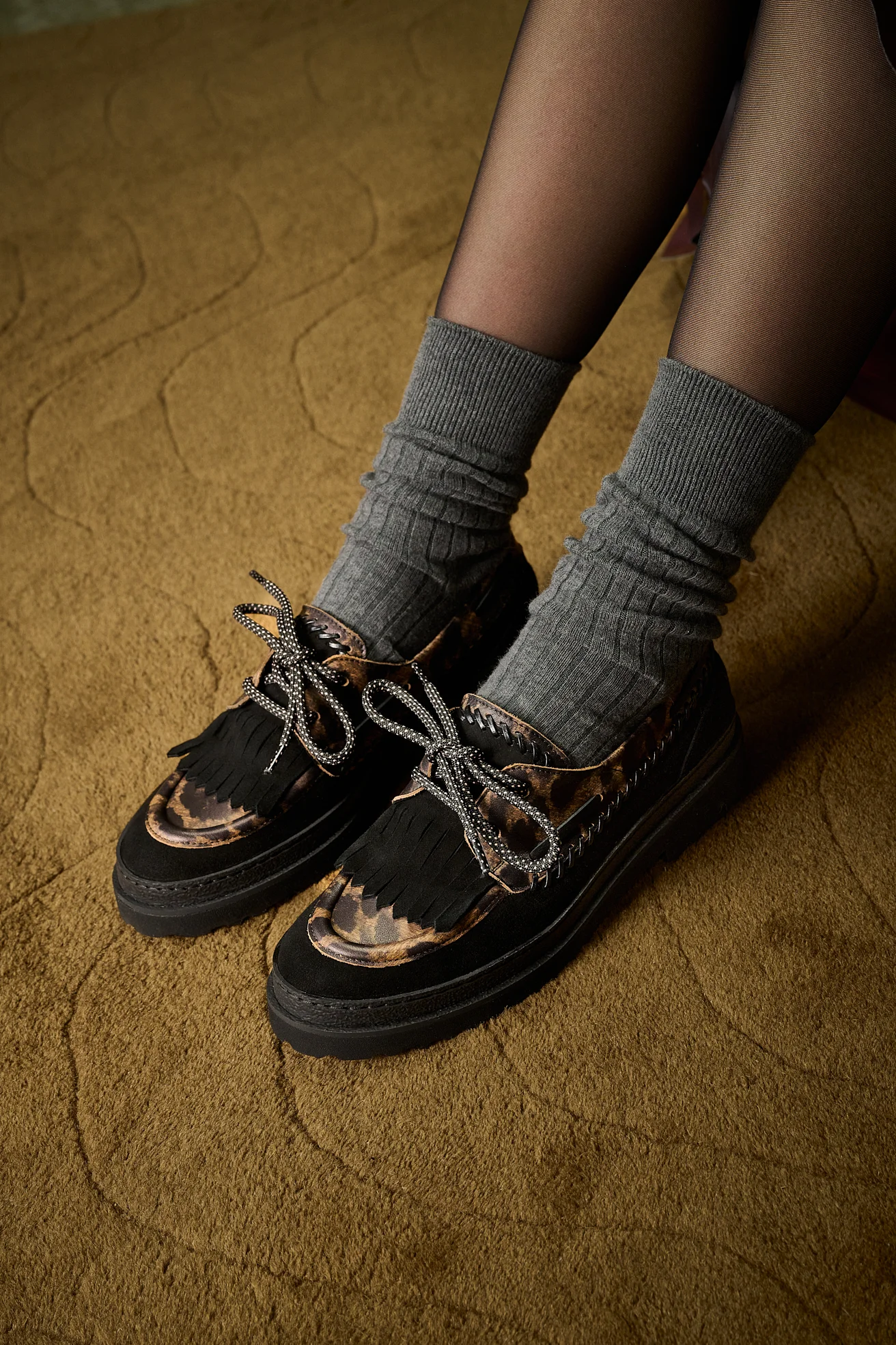 Black suede fringe loafers with leopard-print panels and laces, worn with grey ribbed socks