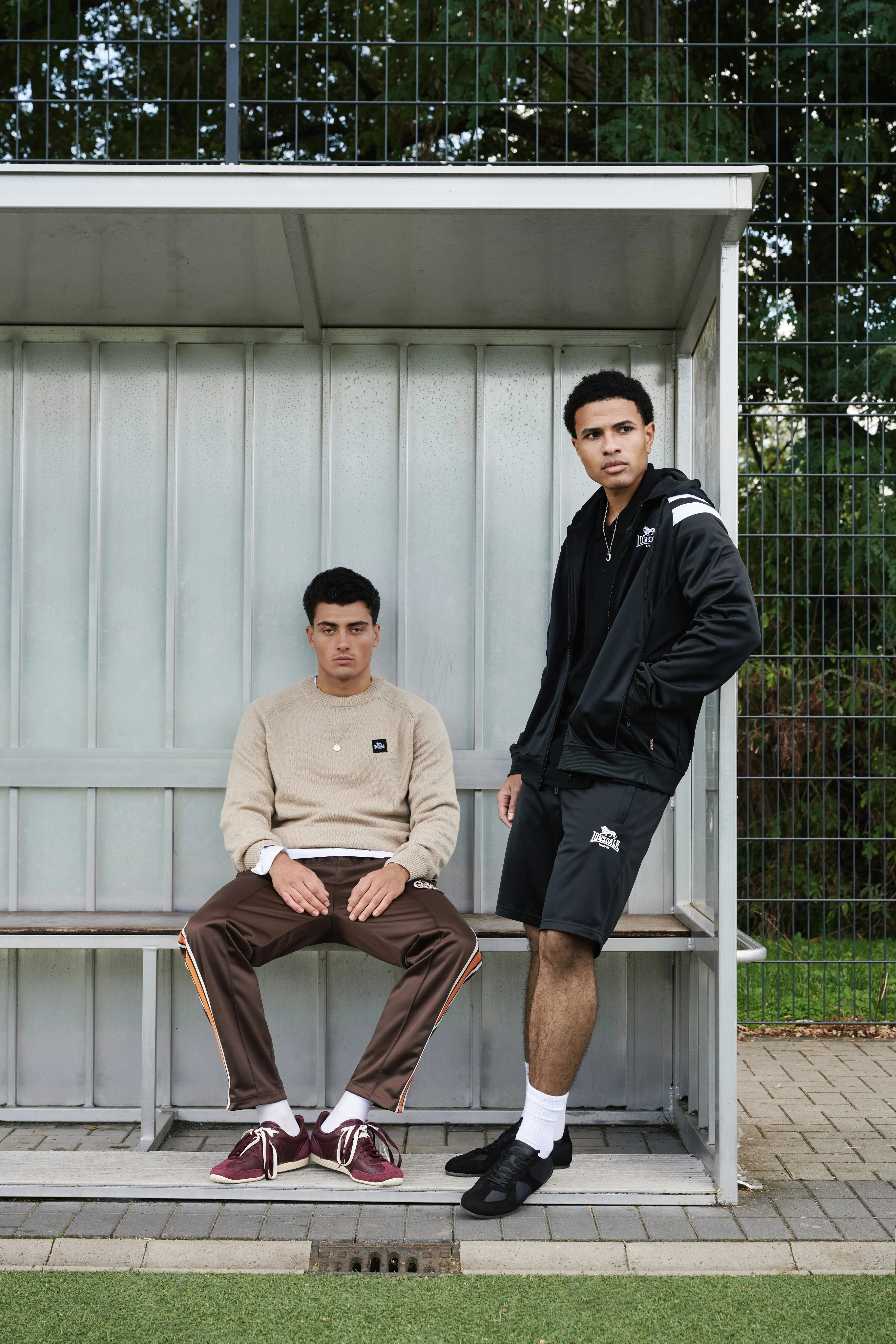 Two men in a dugout by the pitch, one seated in a beige sweatshirt, one standing in a black jacket and shorts