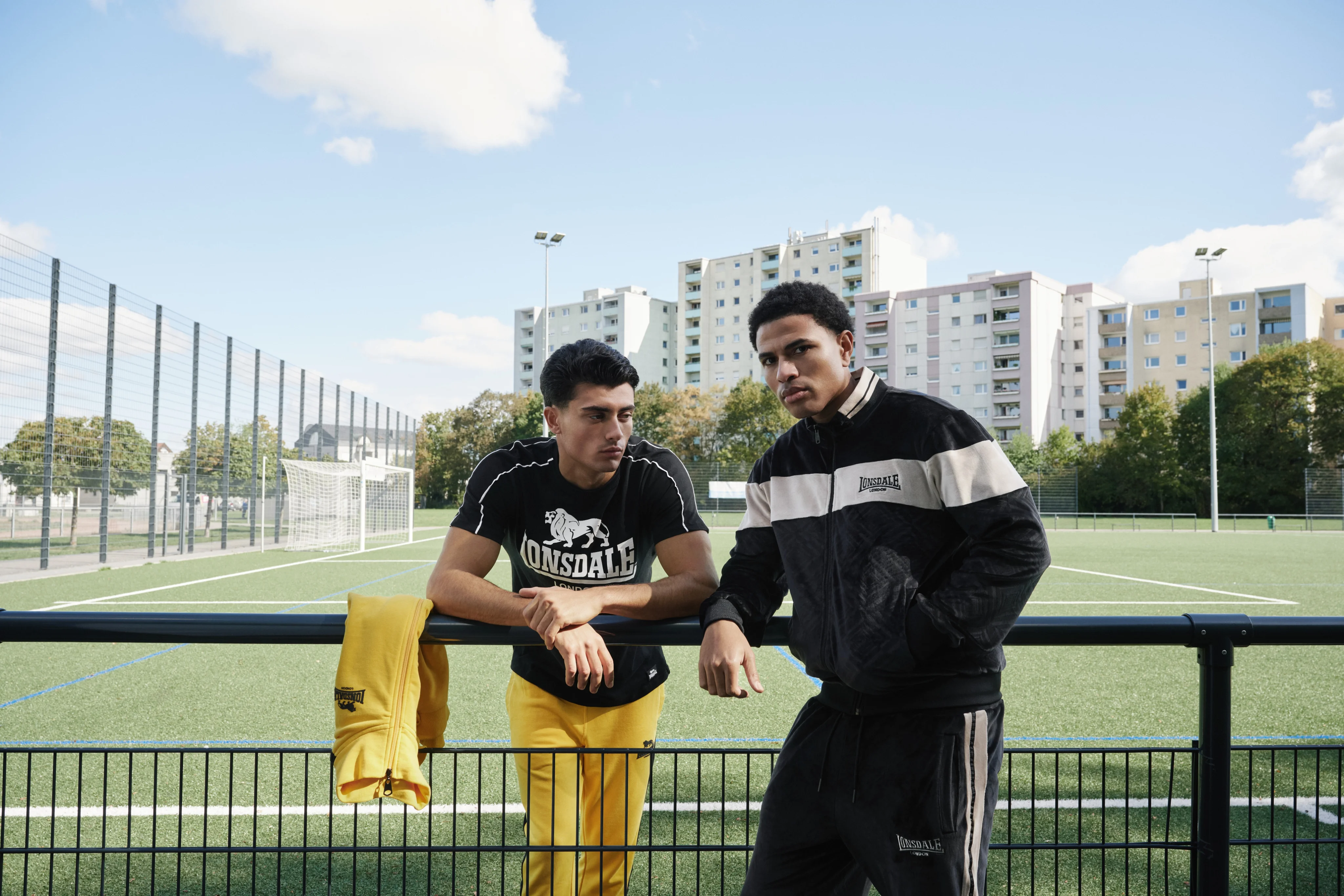 Two men leaning on the railing of a football pitch, apartment blocks behind