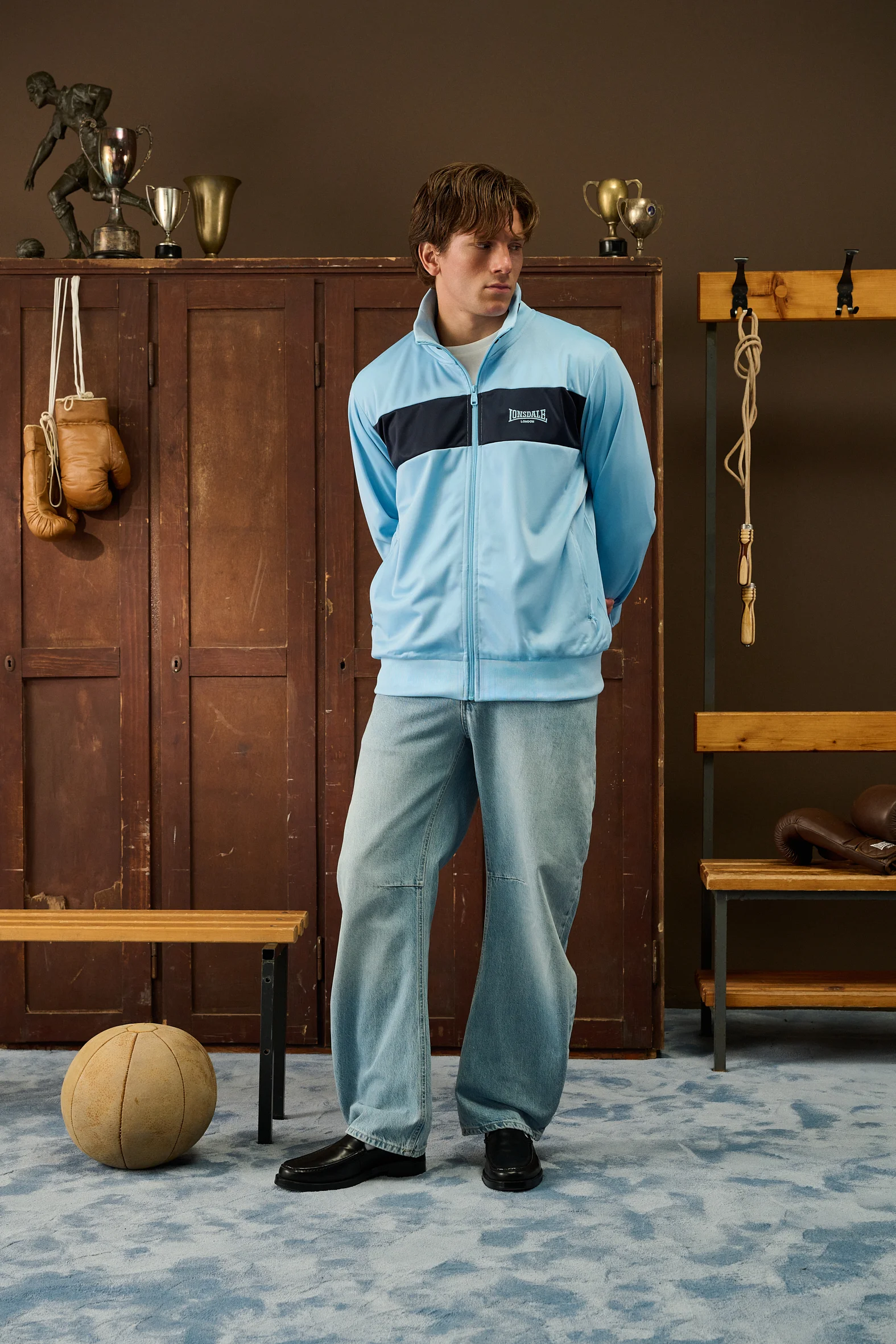 Man in a light blue Lonsdale track jacket and jeans in front of wooden lockers