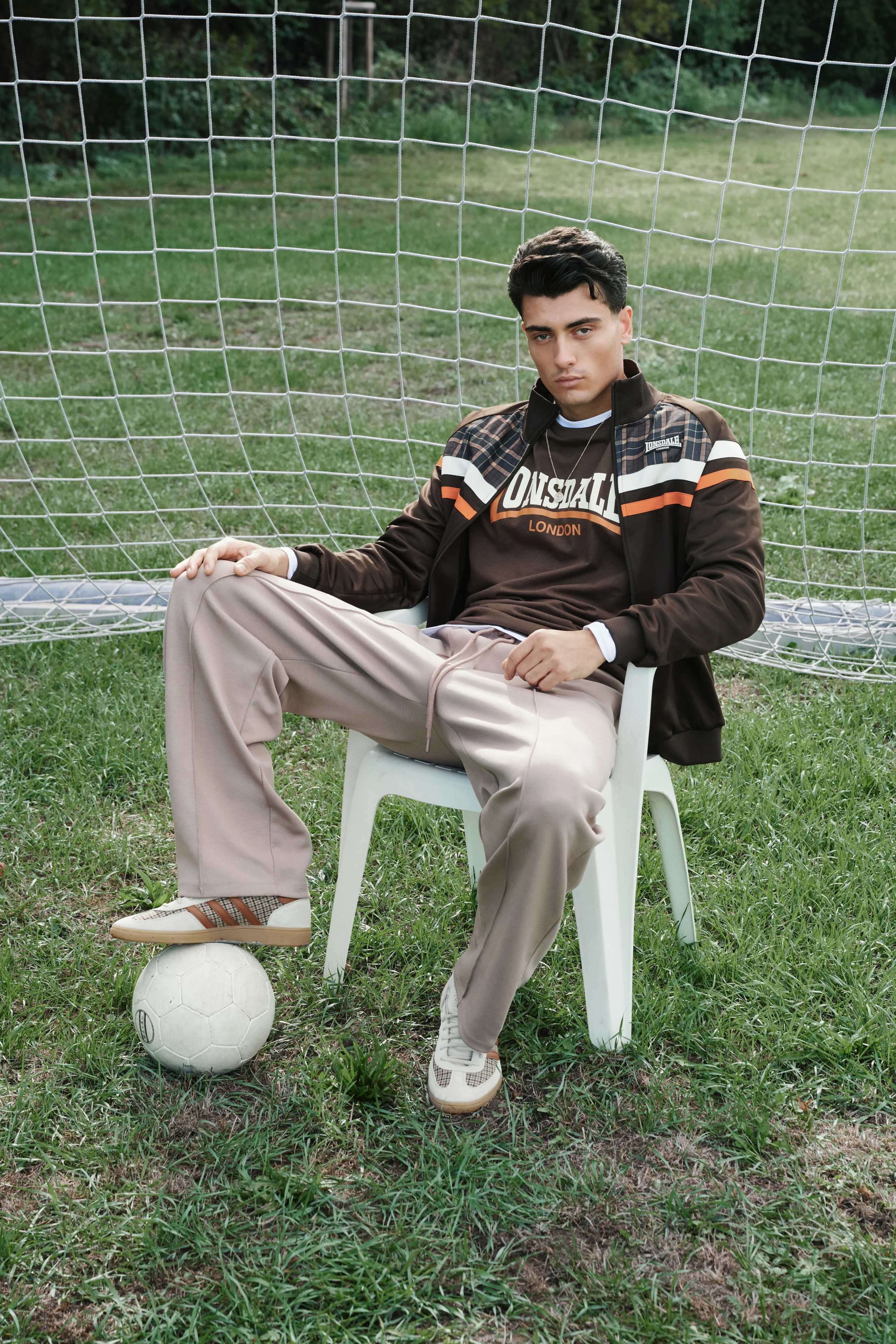 Man in a brown Lonsdale track jacket on a plastic chair, a football at his feet