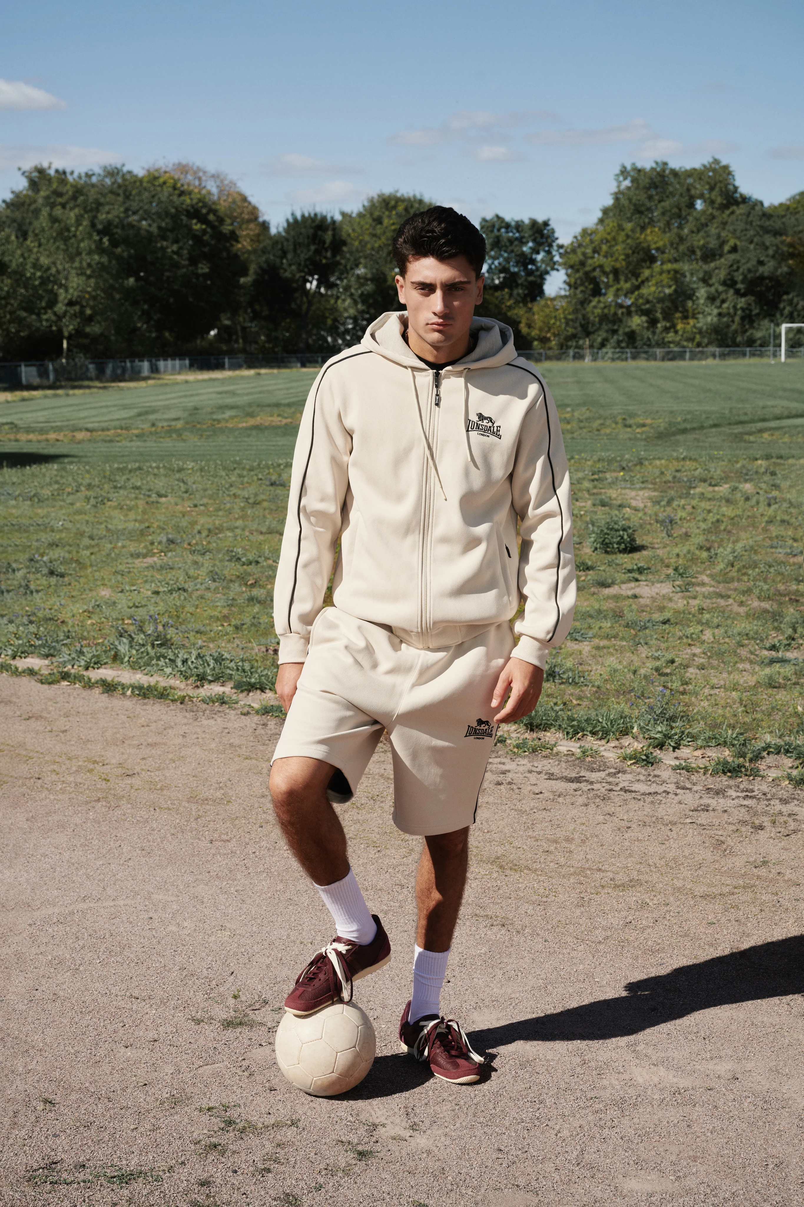 Man in a cream Lonsdale hoodie and shorts with a football on a dirt pitch
