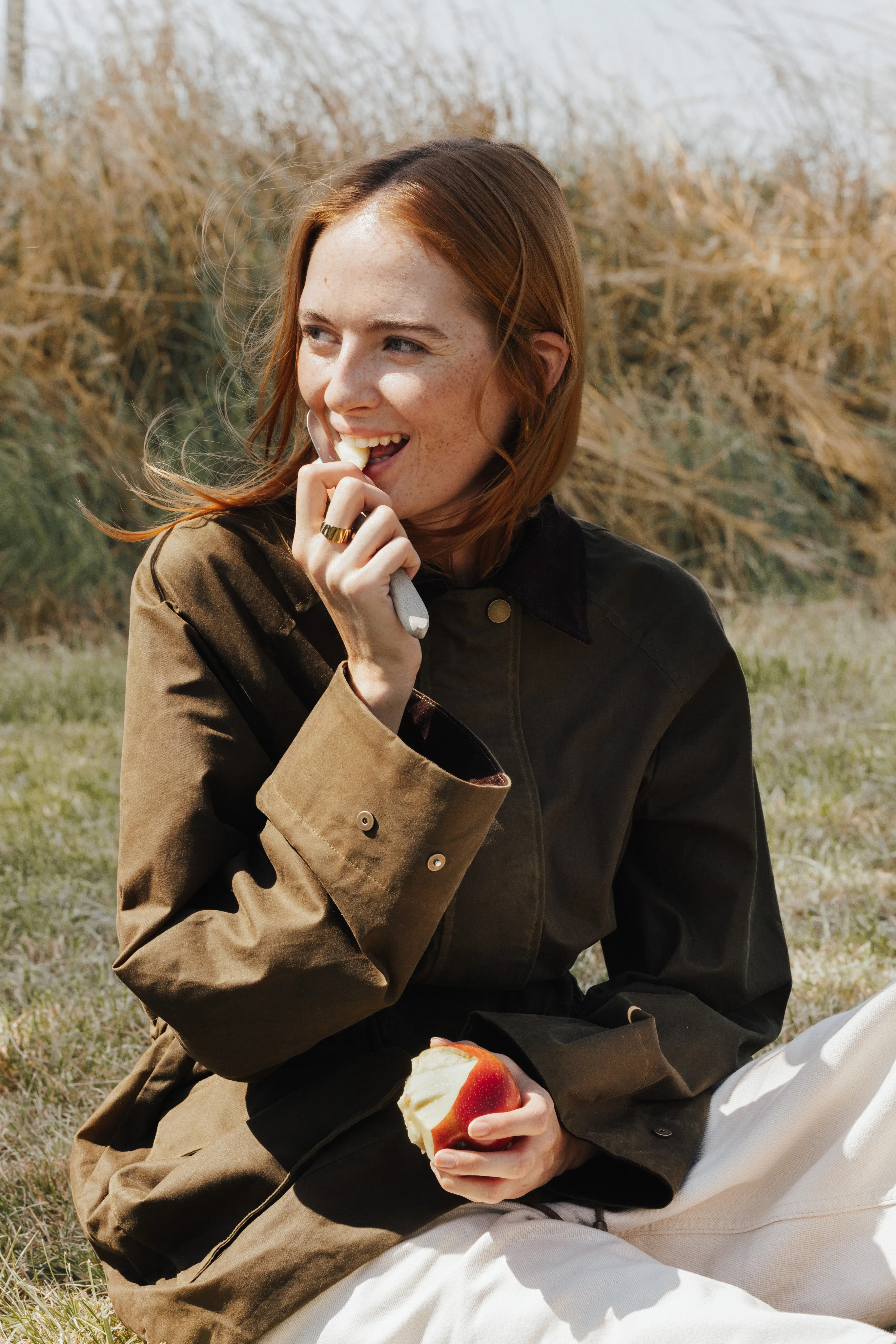 Model in an olive jacket eating an apple in tall grass