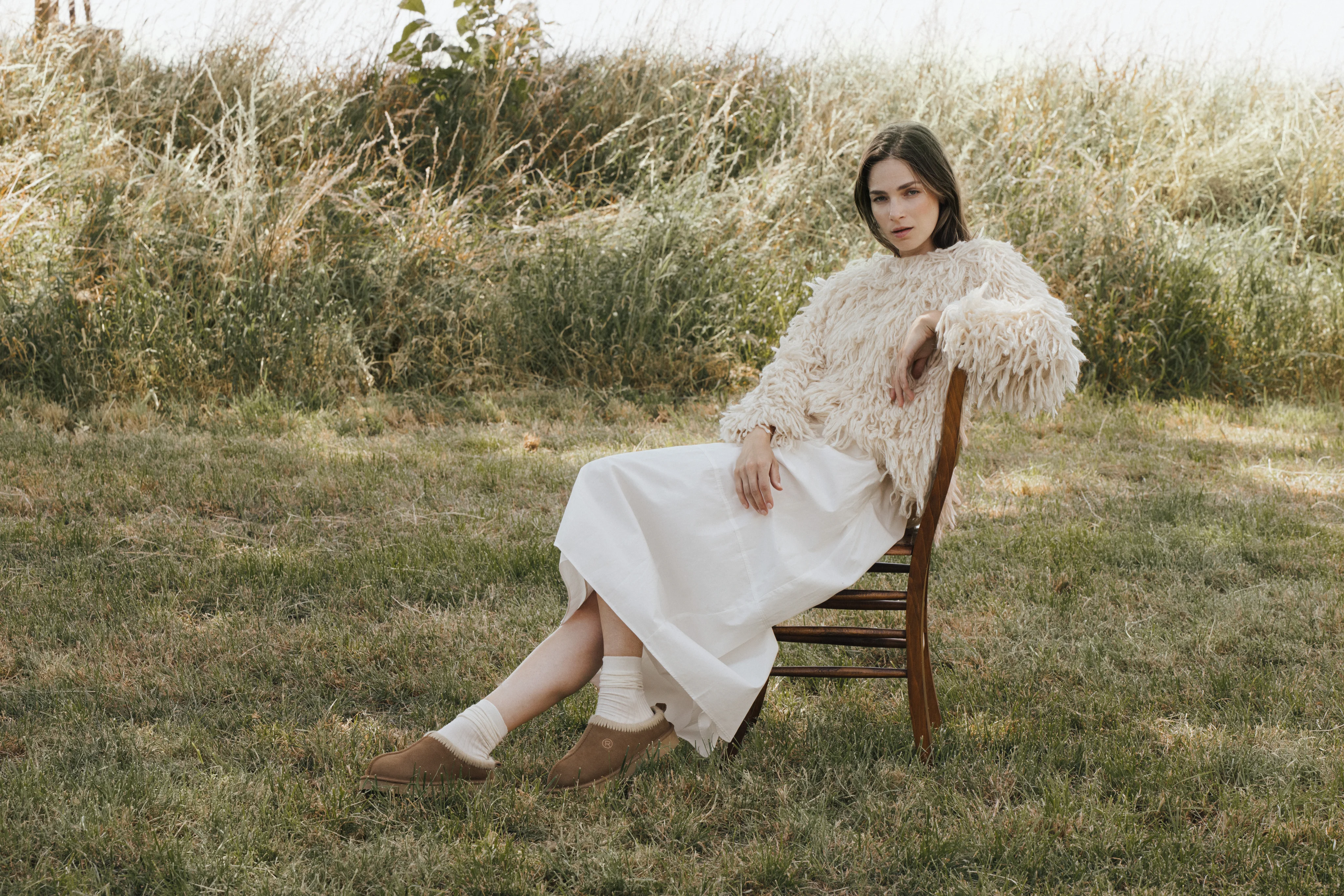 Model in a cream knit and white skirt sitting on a chair in a meadow