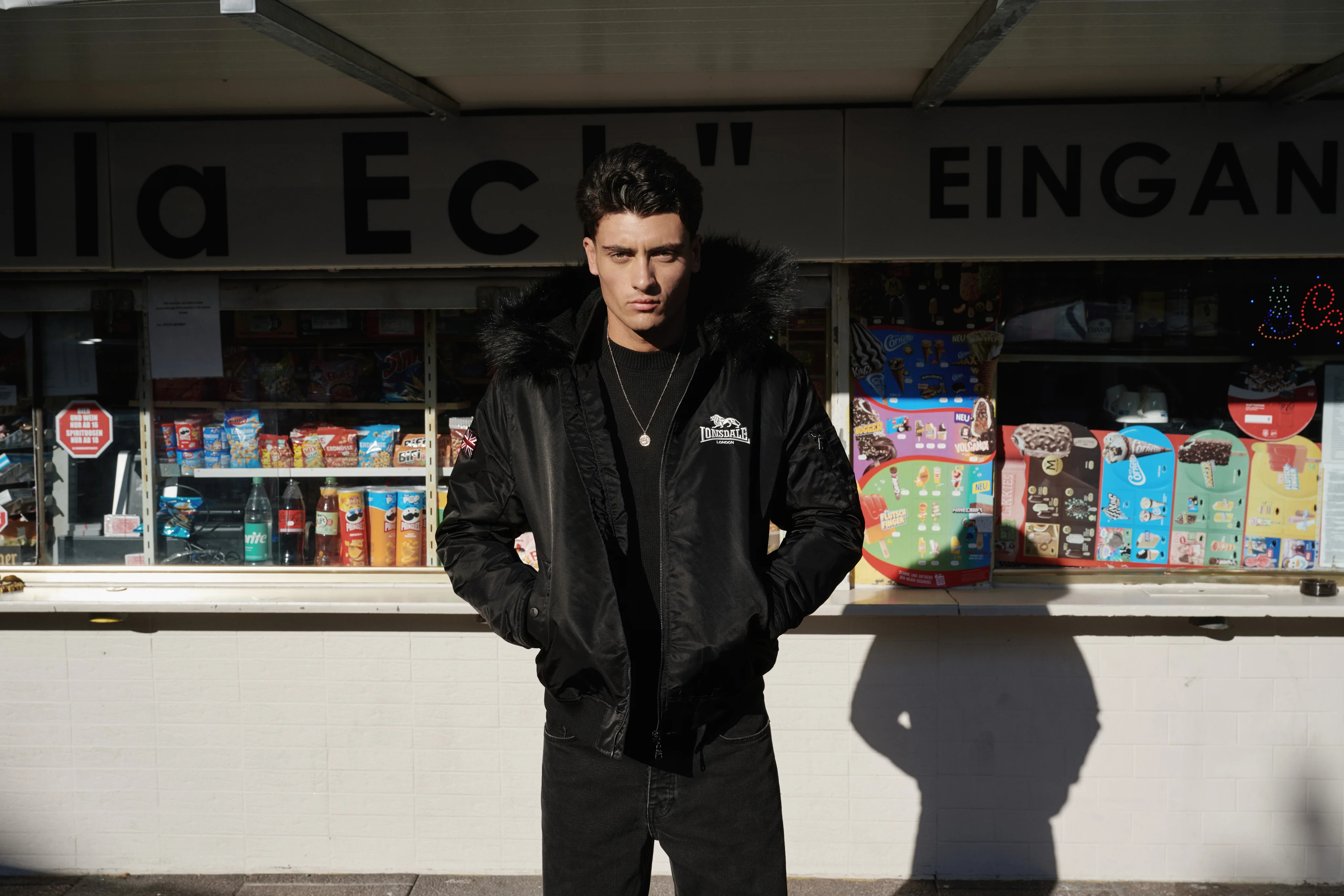 Man in a black Lonsdale bomber jacket in front of a kiosk