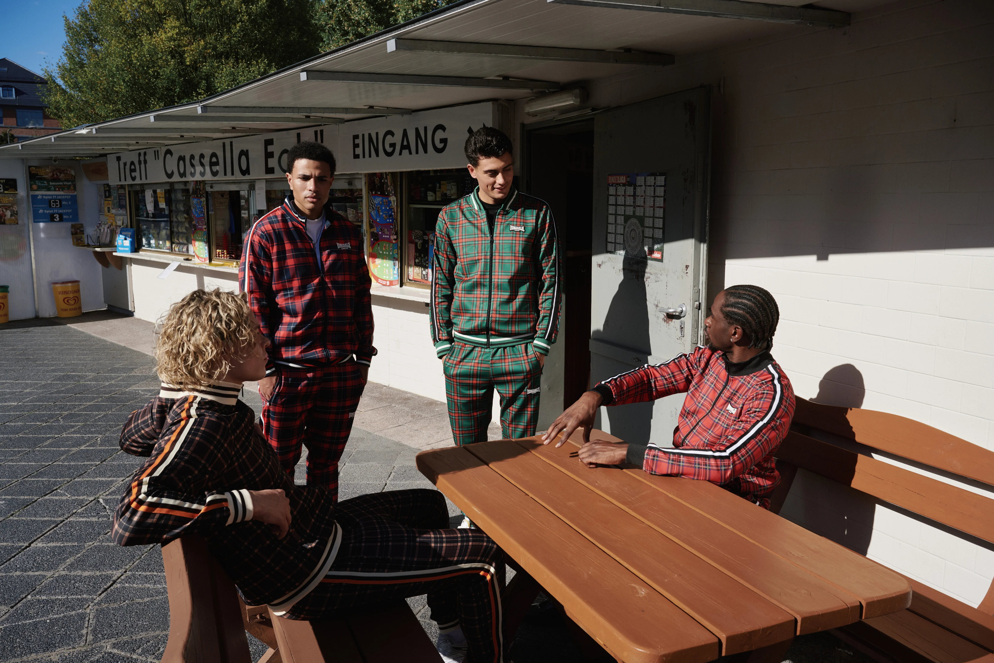 Four men in checked Lonsdale tracksuits at a picnic table in front of a kiosk
