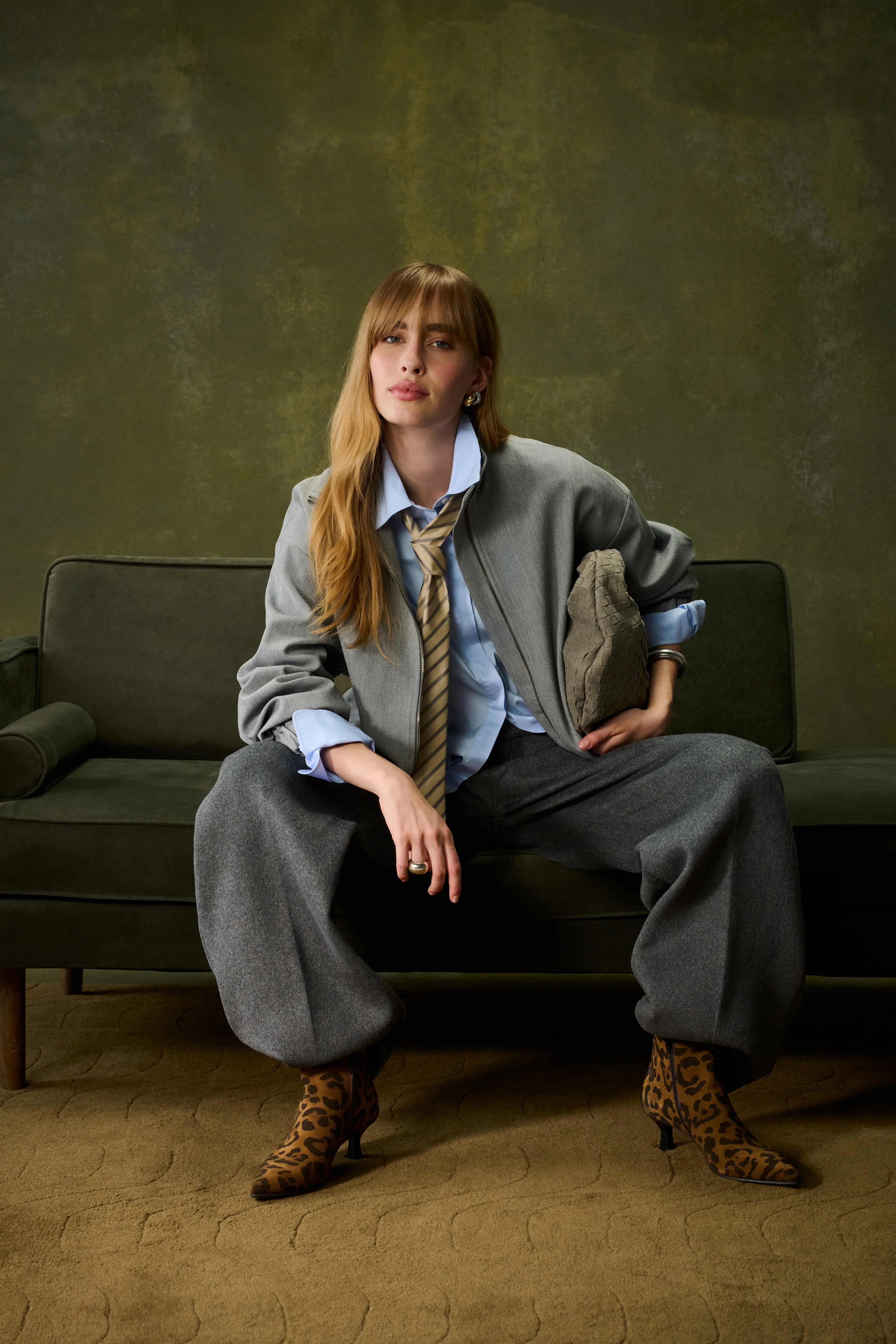Model in a grey jacket, shirt and tie sitting on a sofa in leopard-print ankle boots