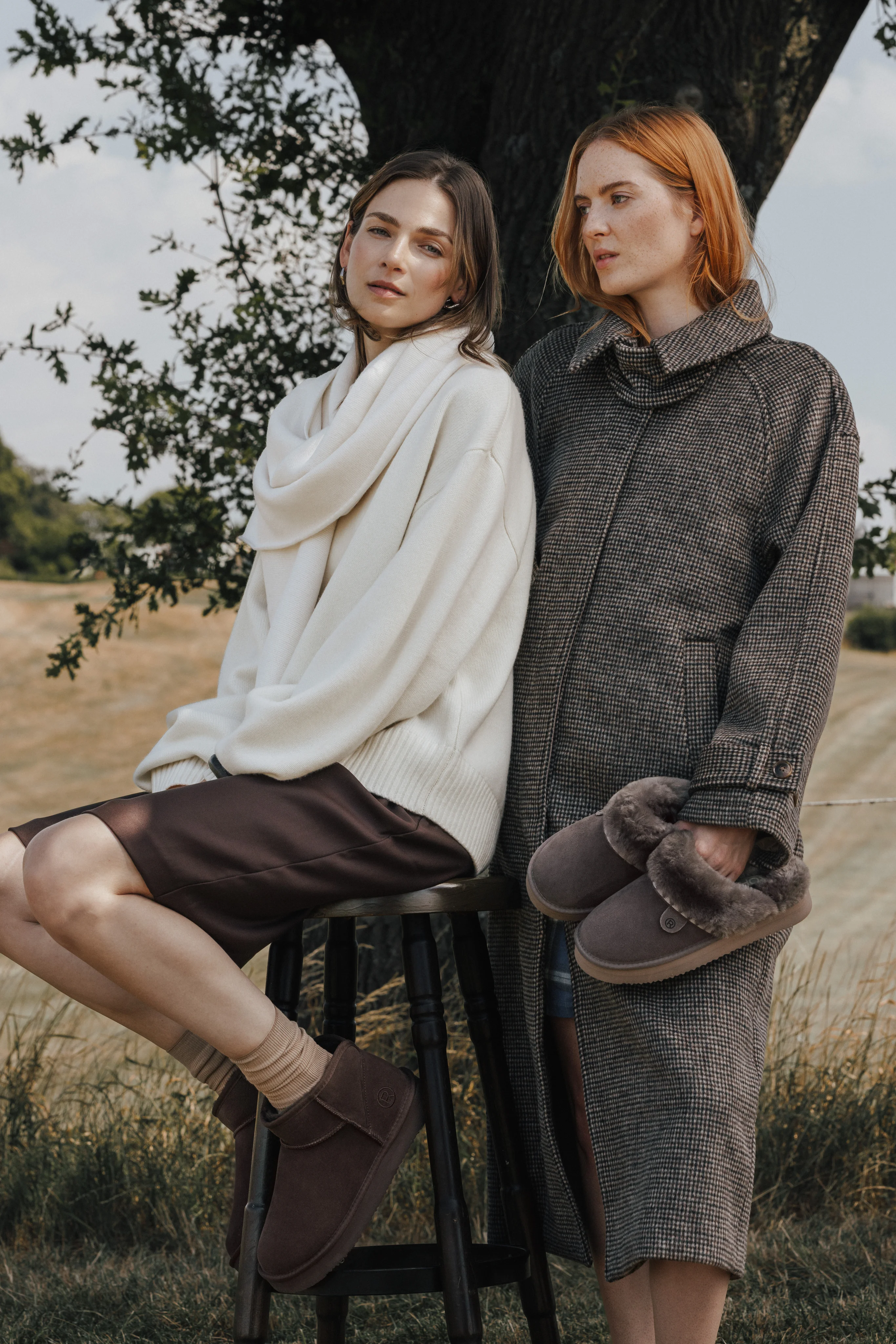 Two models in knitwear and coats holding slippers under a tree