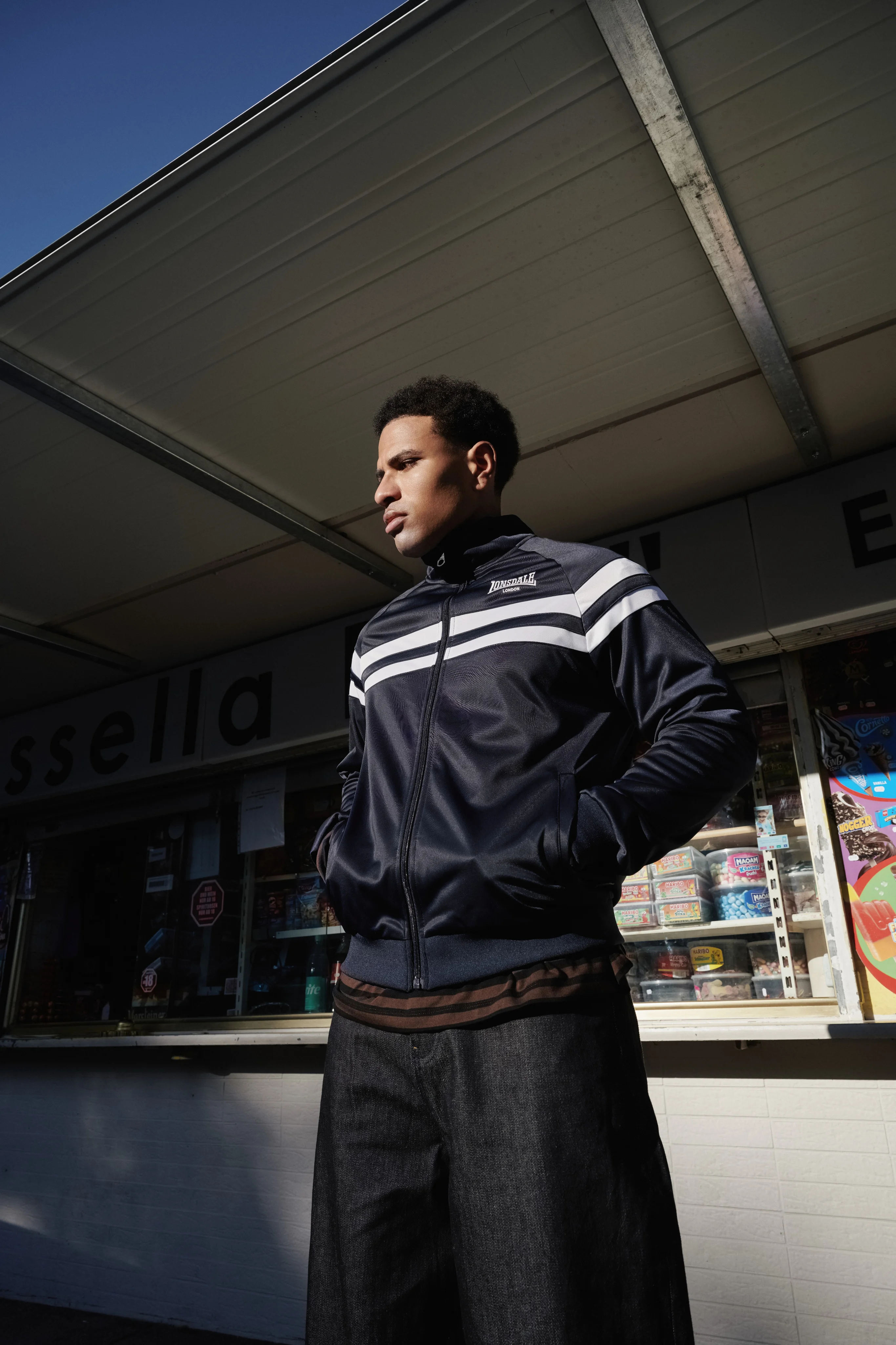 Man in a navy Lonsdale track jacket with white stripes in front of a kiosk