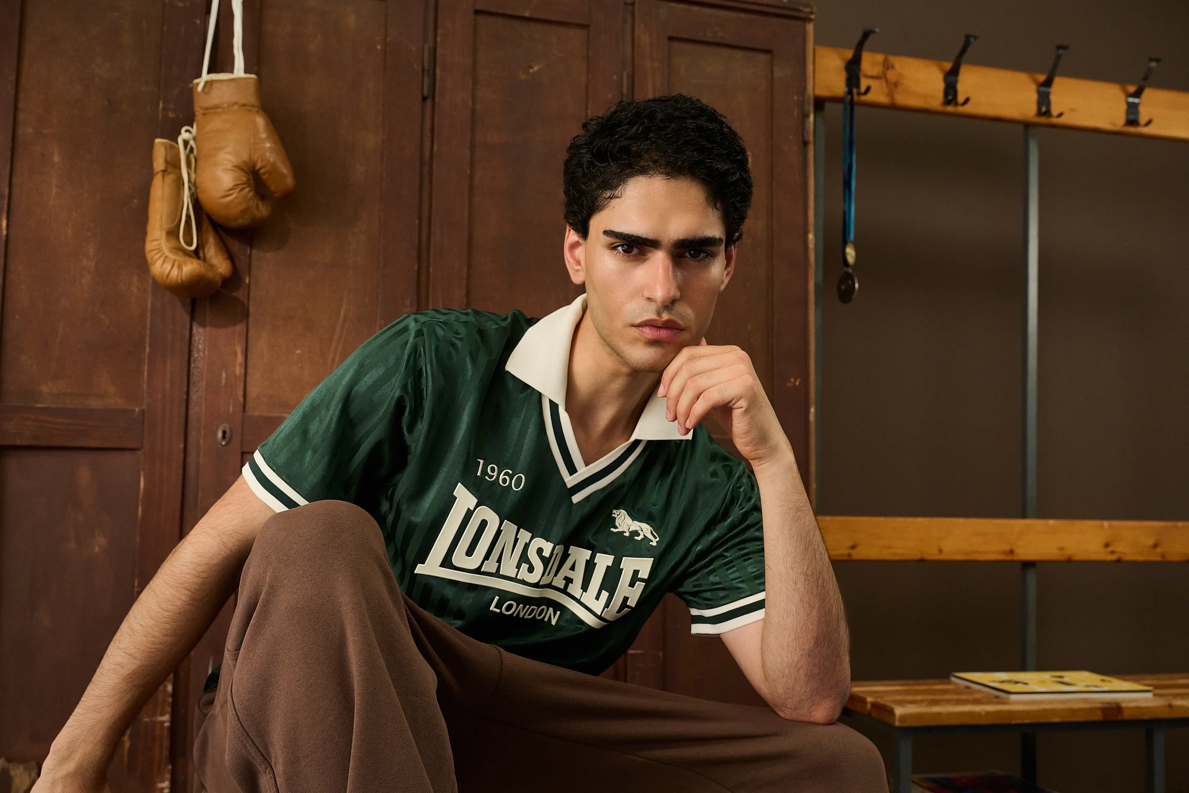 Man in a green Lonsdale shirt with the year 1960 in front of wooden lockers, boxing gloves on a hook