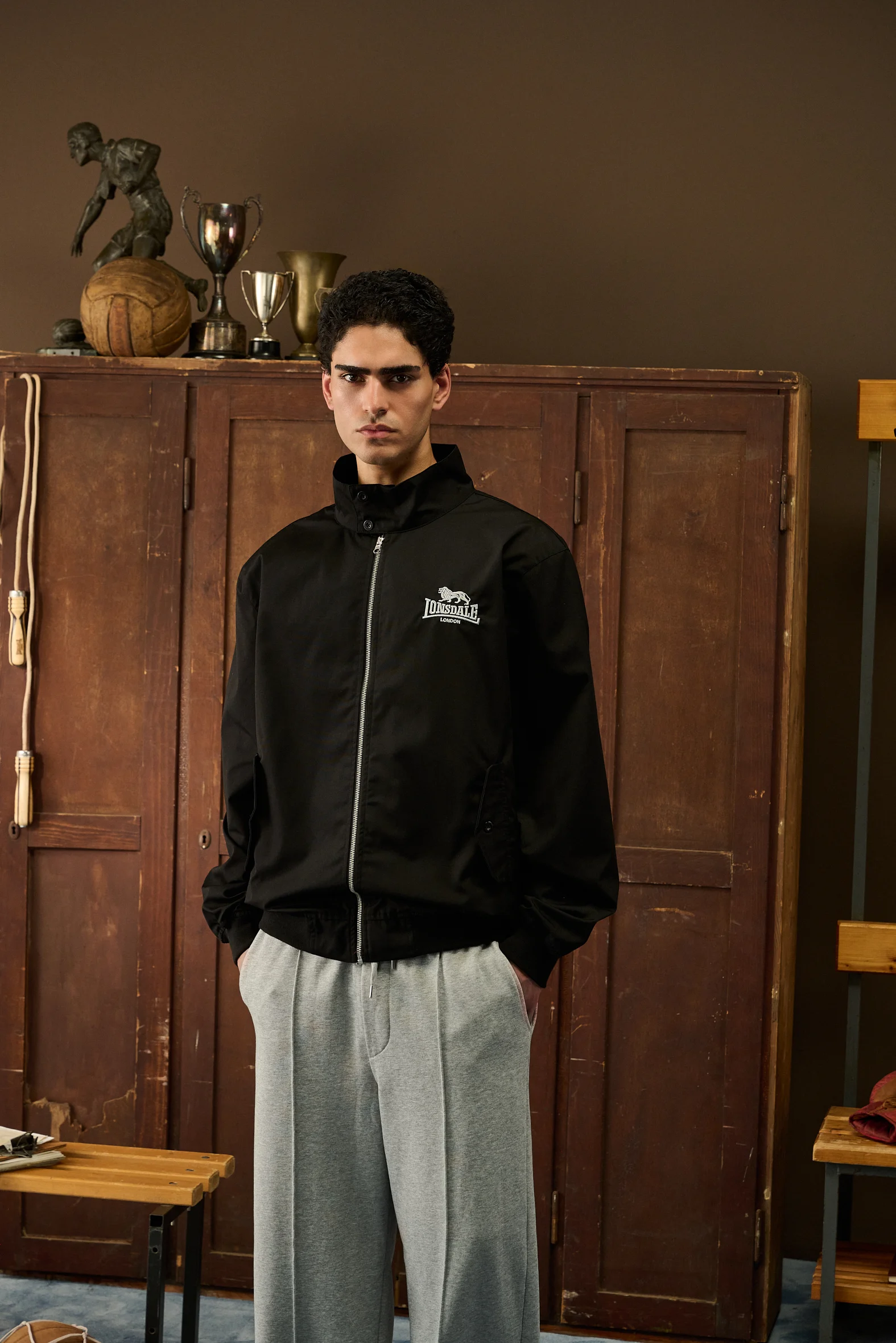 Man in a black Lonsdale track jacket and grey trousers in front of wooden lockers