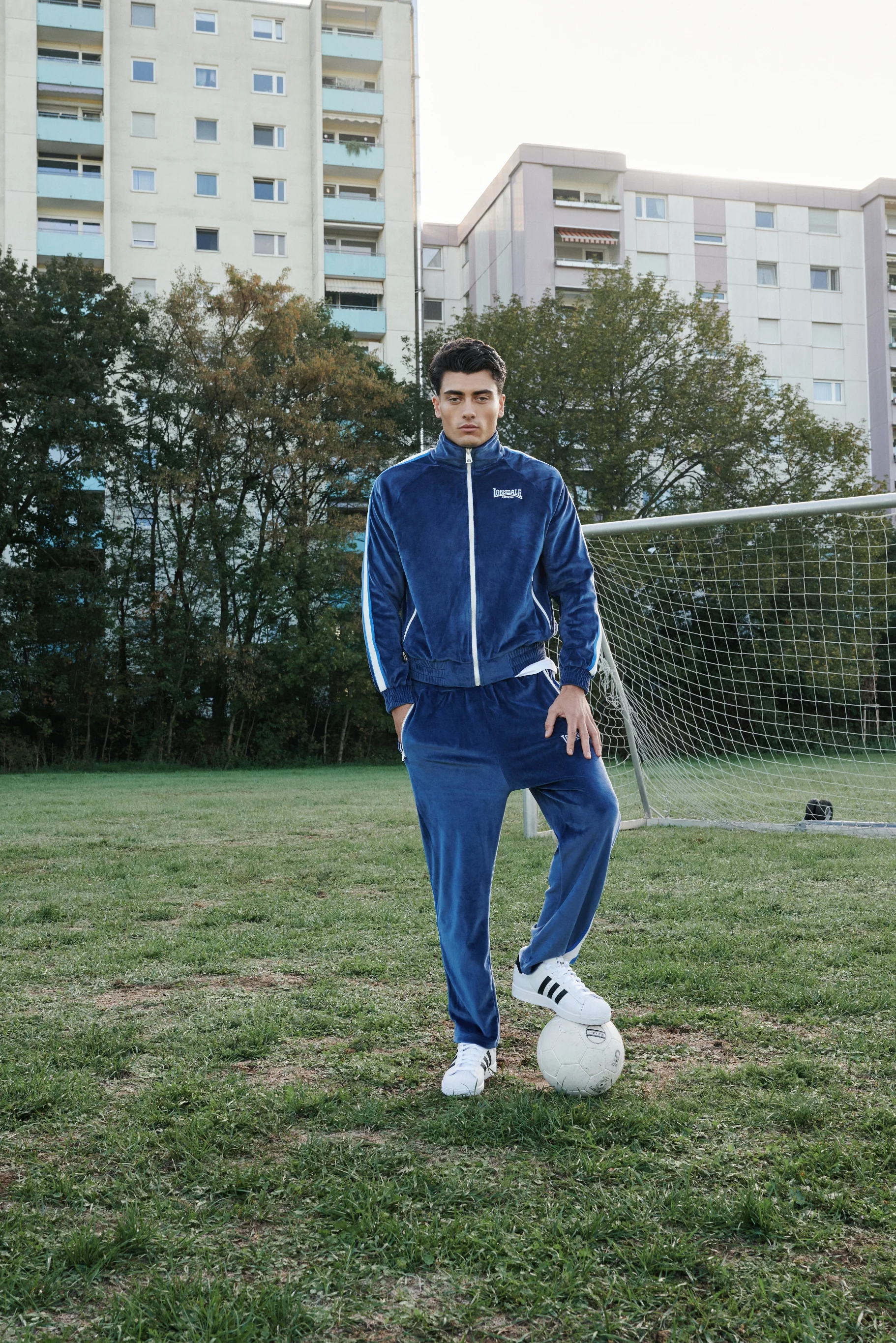 Man in a blue Lonsdale tracksuit with one foot on a football in front of a goal