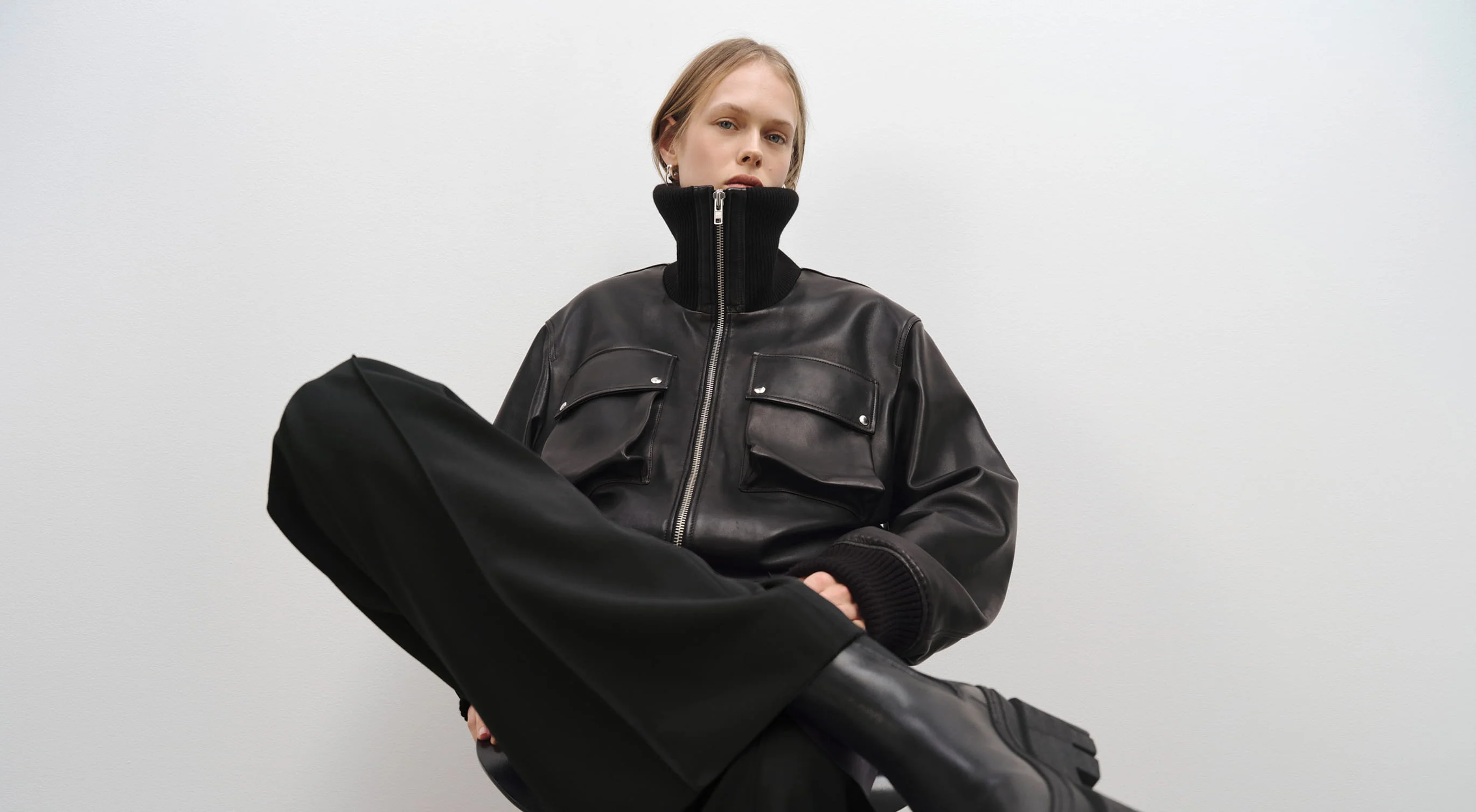 Model in a black leather jacket and wide black trousers sitting against a white wall.
