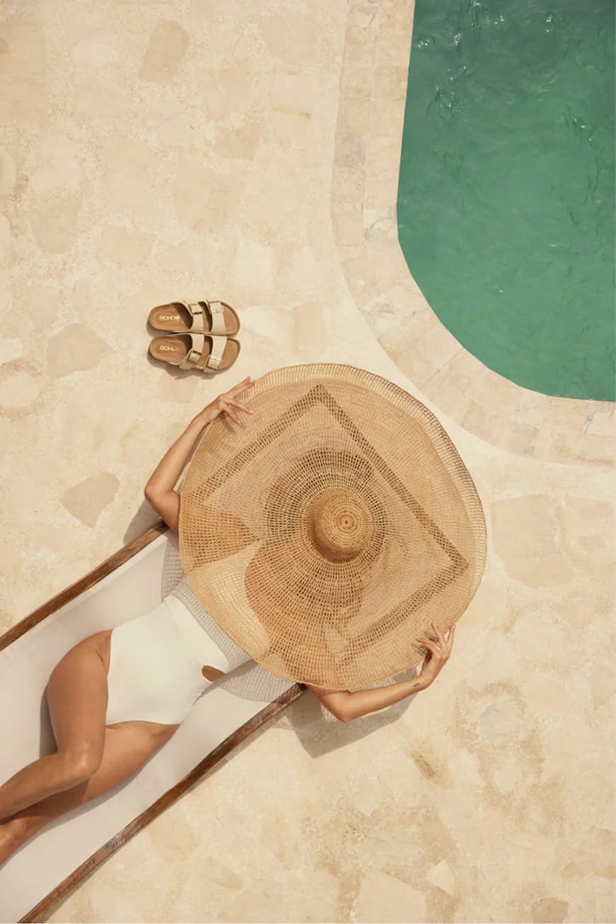 Woman in a white swimsuit lying under a straw hat, sandals beside her.