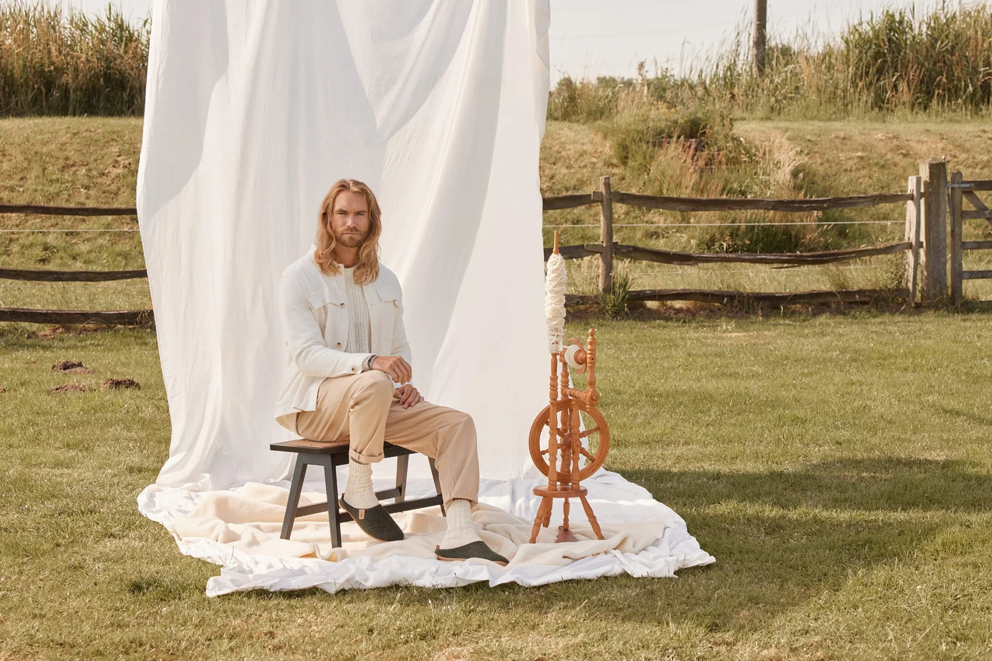 Man with long blond hair sitting next to a spinning wheel in a meadow.