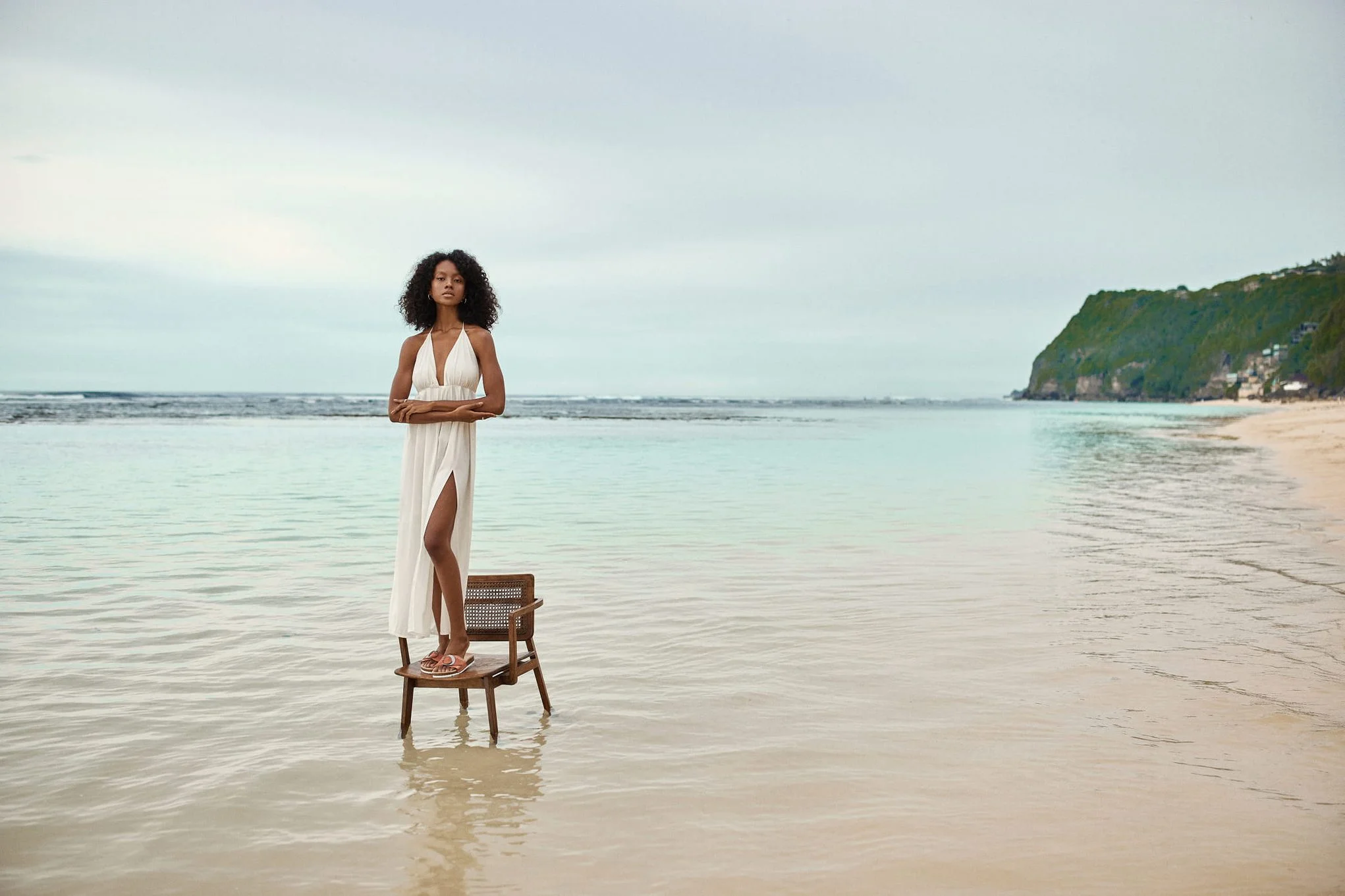 Woman in a white dress standing in shallow sea water next to a chair.