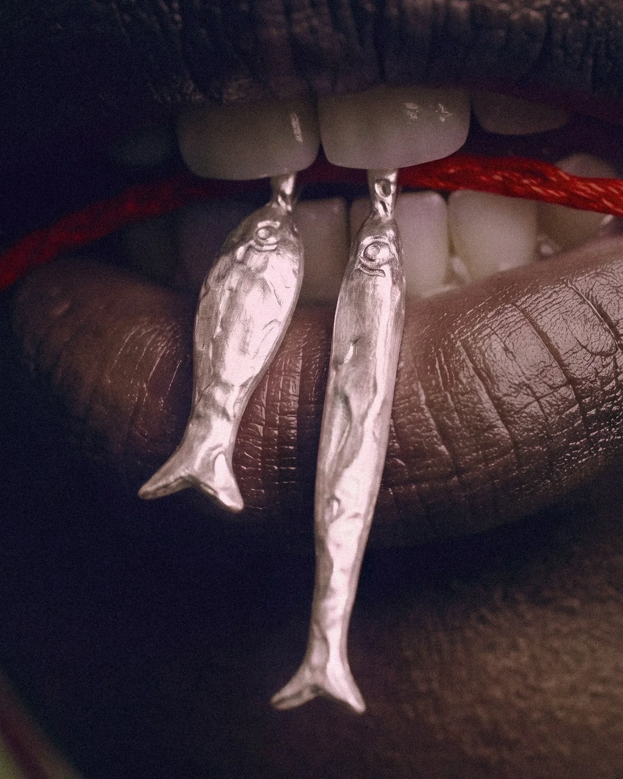 Close-up of a mouth holding silver fish pendants on a red cord