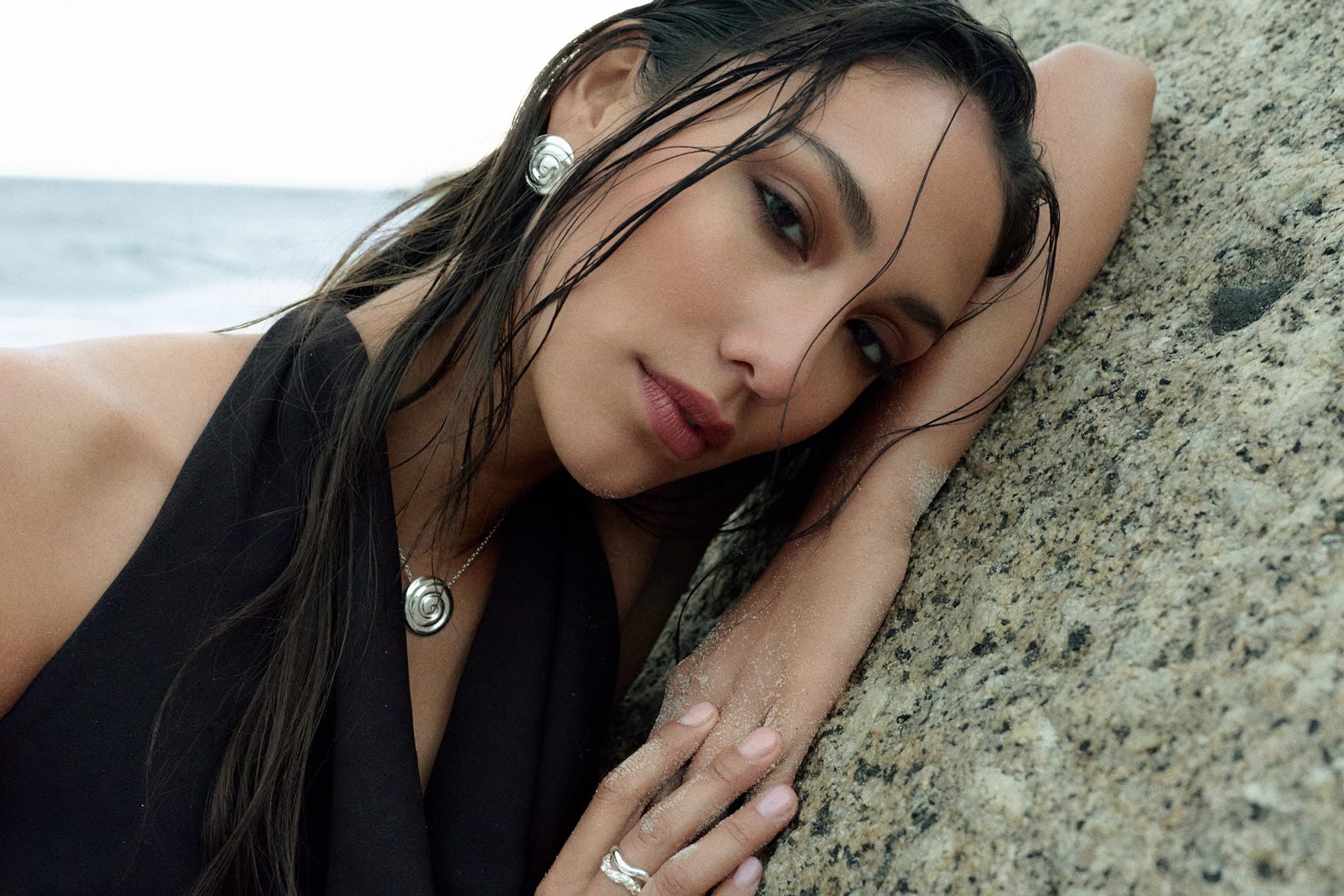 Woman in black leans on a rock at the beach, wearing jewelry.