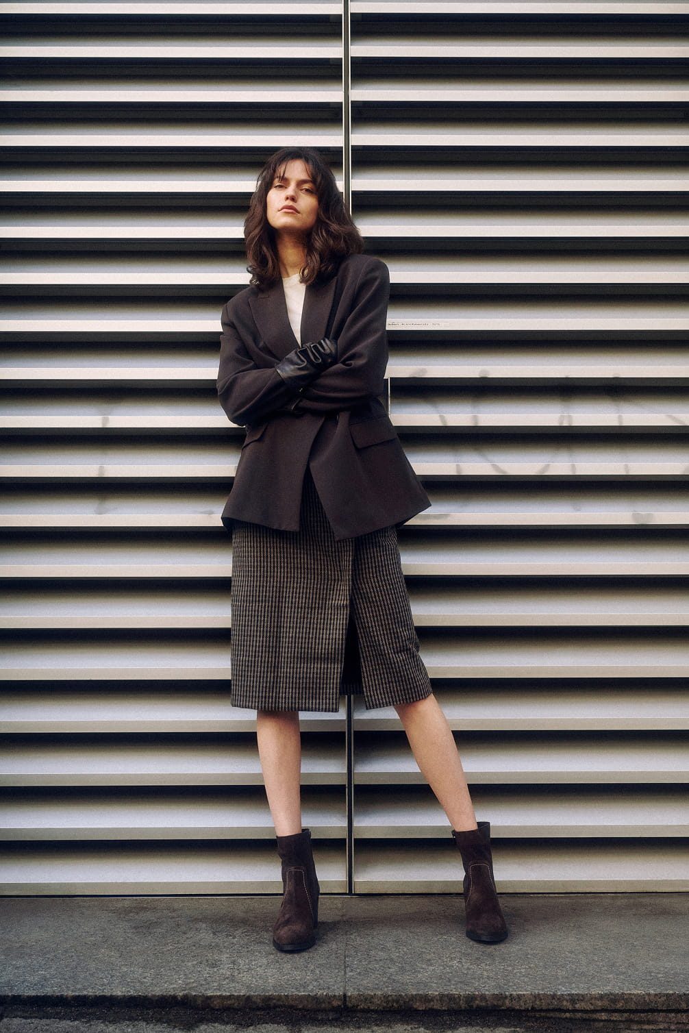 Woman in dark blazer and plaid skirt posing against wall