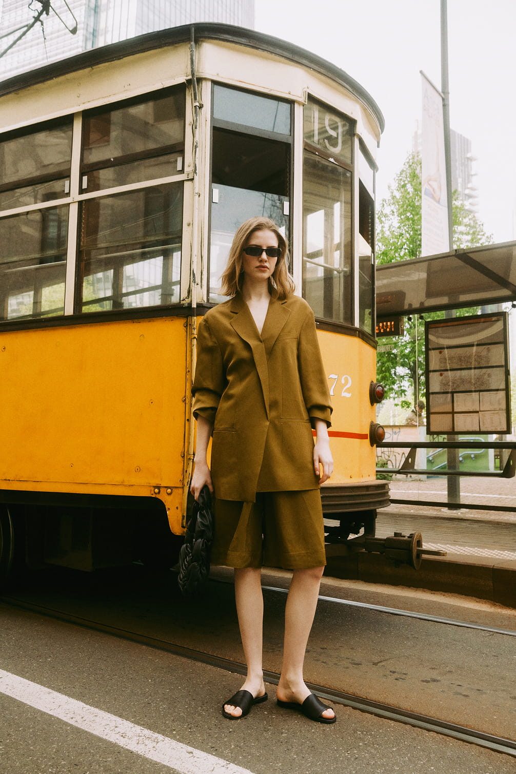 Woman in olive suit stands by yellow tram.
