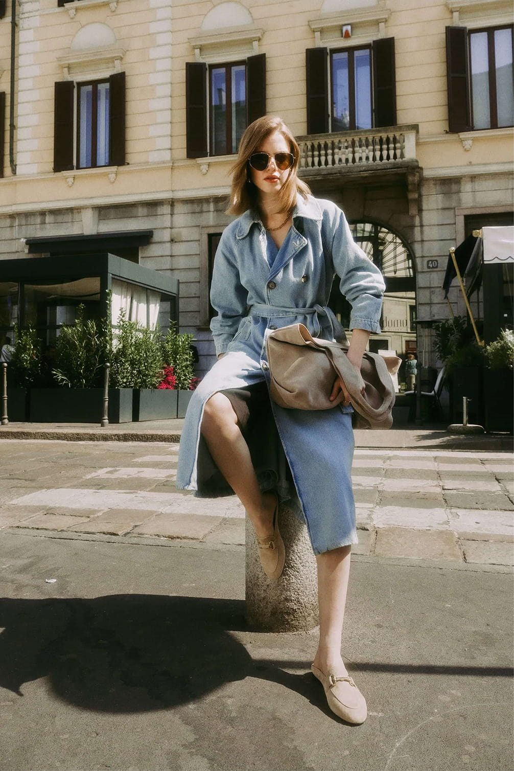 Woman in denim trench coat sits outside a building.