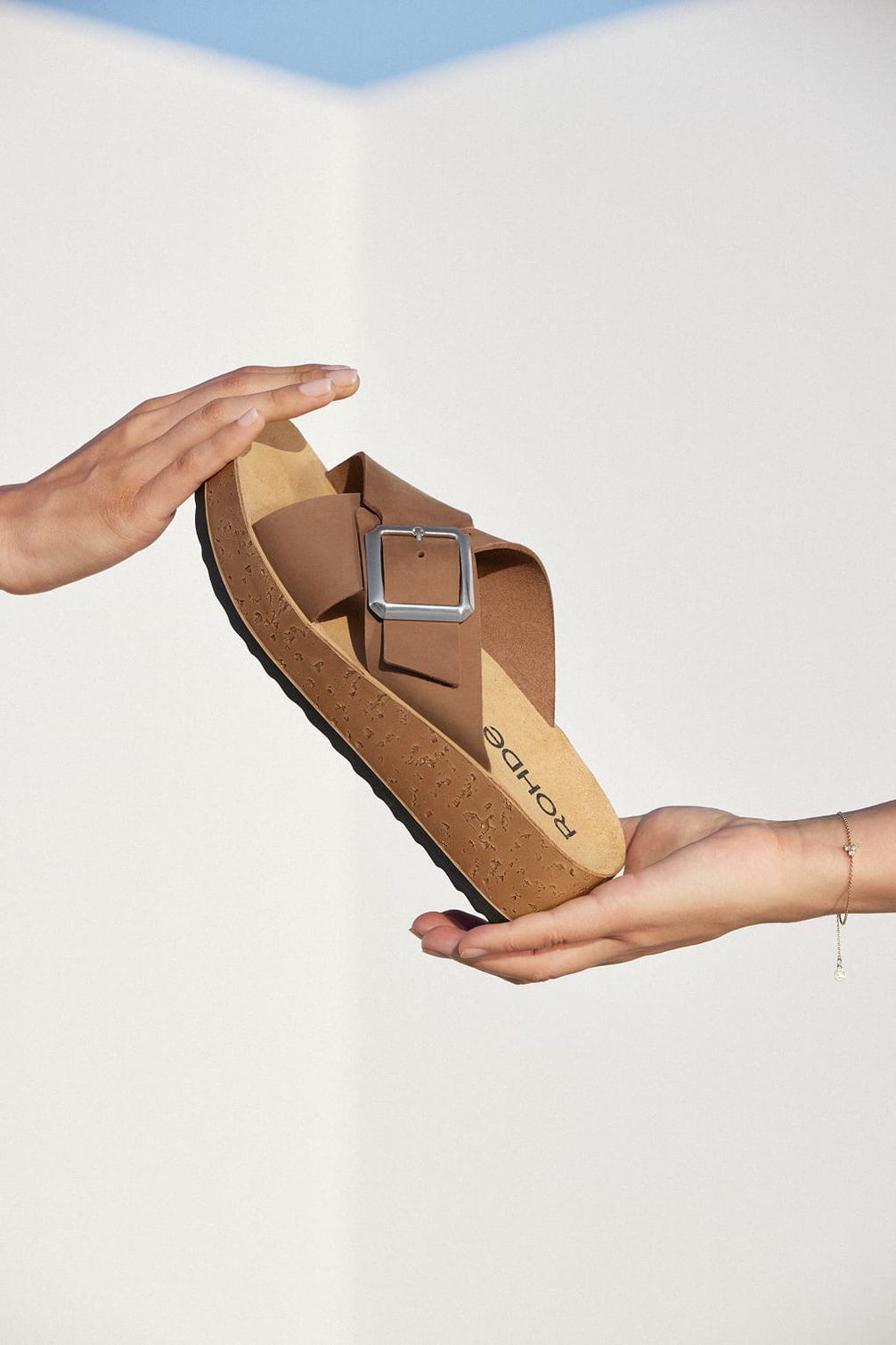 Brown sandal with silver buckle, presented by hands.