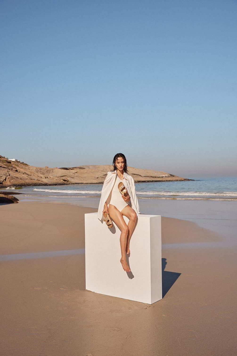 Woman in white swimsuit on beach with sandals