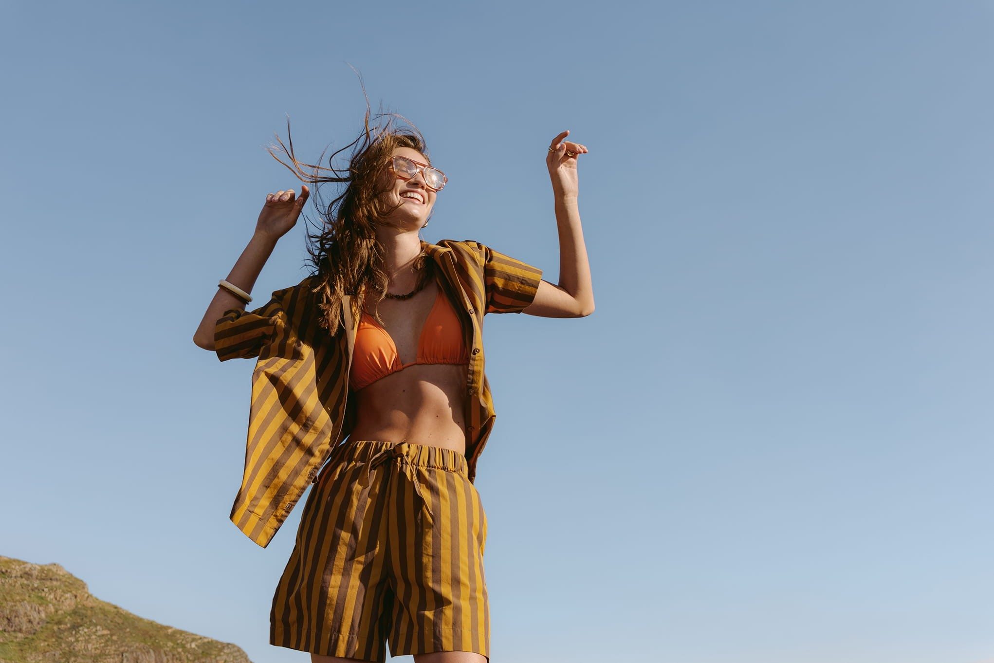 Woman in striped outfit raises hands against clear sky.