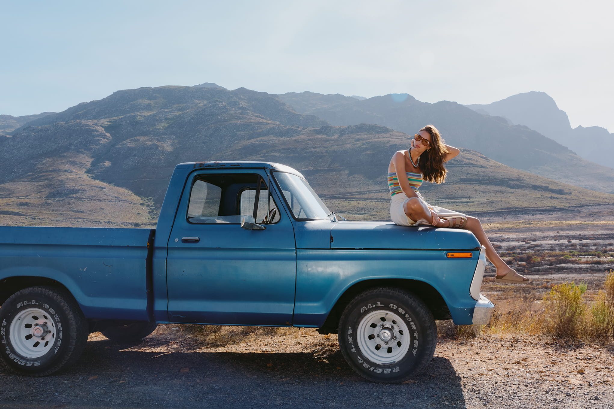 Woman sitting on a blue vintage truck in a dry, mountainous landscape.