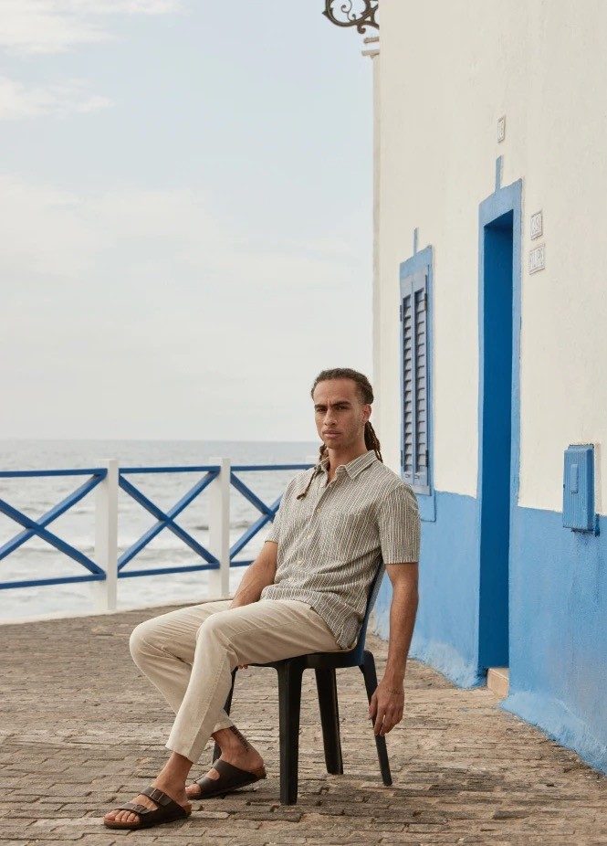 Man in striped shirt and pants sitting on chair near blue and white building.