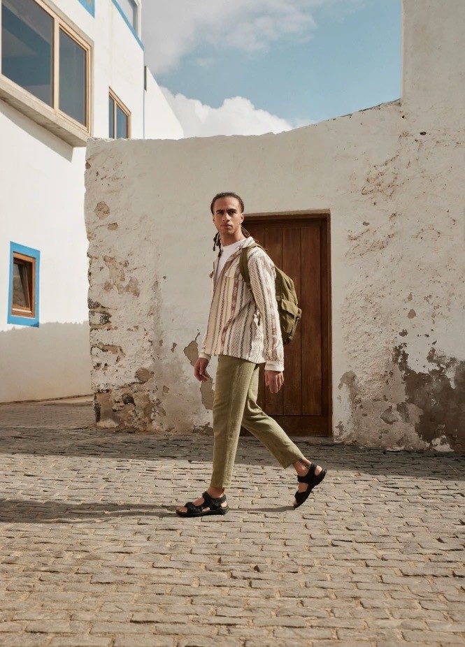 Man with backpack walks down cobblestone street in neutral outfit.